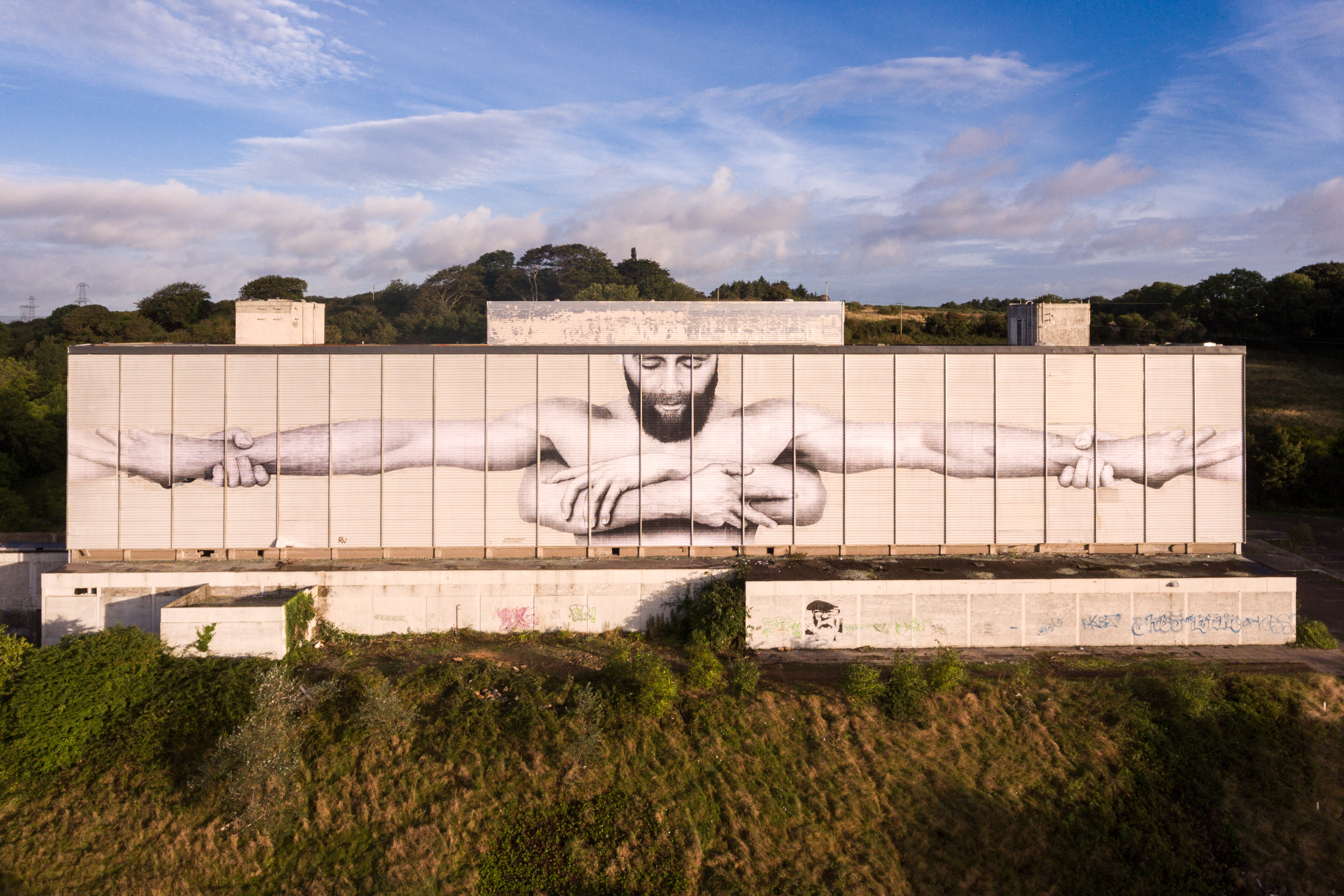 Joe Caslin's new mural on the back of a hotel in waterford showing a bearded man holding arms around him and supported by another pair of arms from behind