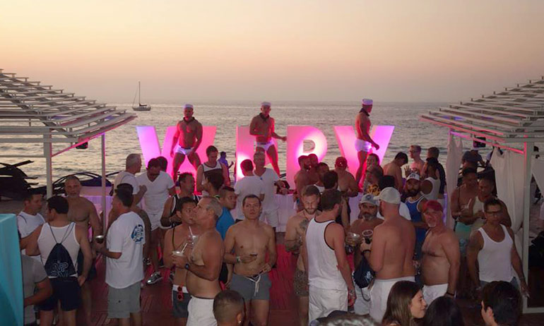 A party at the White Party Palm Springs with people dancing in front of a pink illuminated sign, one of the top ten queer travel events of 2017