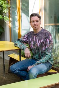 Ray O'Neill smiling on a bench with a top decorated with tulips at the cake cafe and slice which are both owned by Ray O'Neill