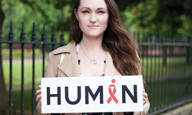 A woman holding a sign with the word Human written on it where the A is a red ribbon to raise awareness about the exhibition taking place in June 2017