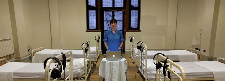 A nurse standing in front of a computer hooked up to headphones on hospital beds as part of Shannon Sickles' Reassembled, Slightly Askew show