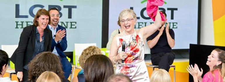 Minister for Children and Youth Affairs holding up a pink flag and a microphone in front of signs for the LGBT helpline, as she speaks about helping LGBT Young People