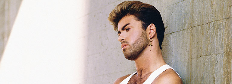 George Michael with a cross earring in a white tank top against a wall