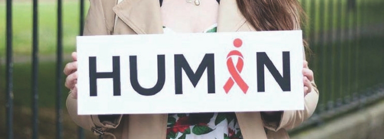 A person holding an all together human sign with a red ribbon in place of the A in Human
