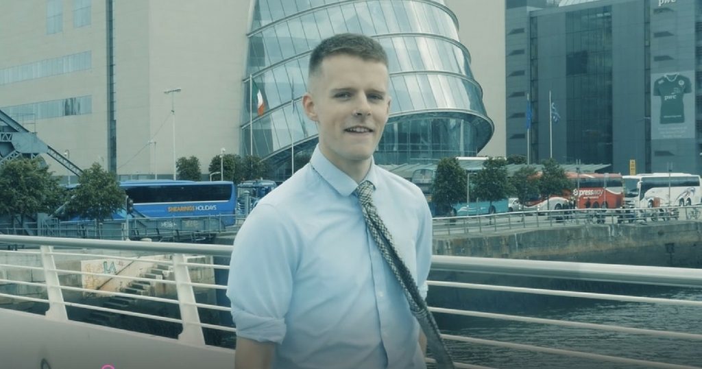 A shot from the newly released video 'Gay, Male, Votes Fine Gael' of Oisin McKenna standing in front of the Dublin Convention Centre performing his poem to camera. He is dressed smartly as a politician, wearing a shirt and tie.