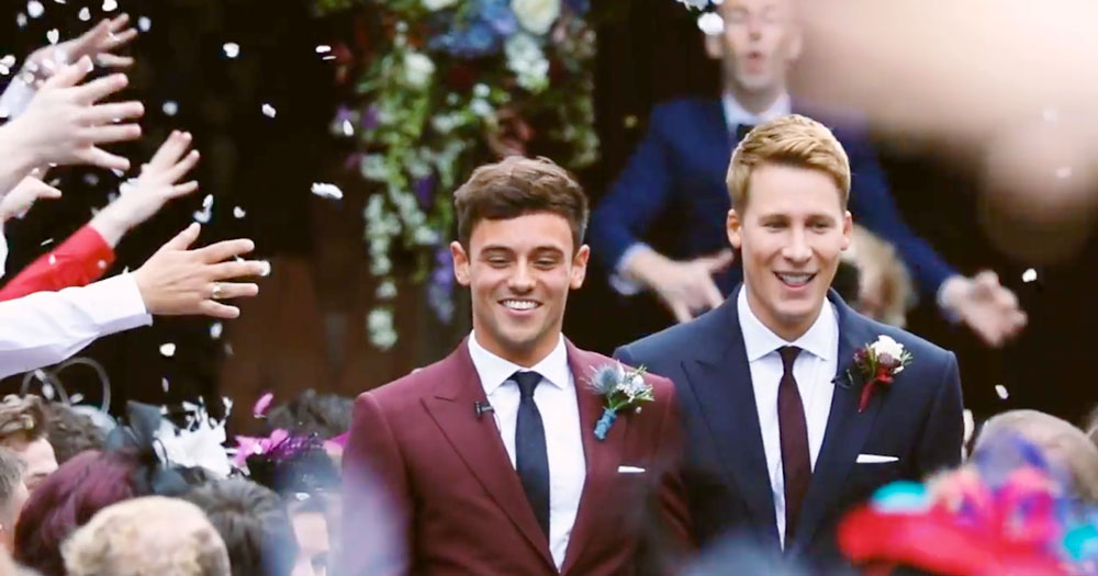 Tom Daley and Dustin Lance Black in a still frame from their wedding video walking in wine and navy suits with confetti in the air