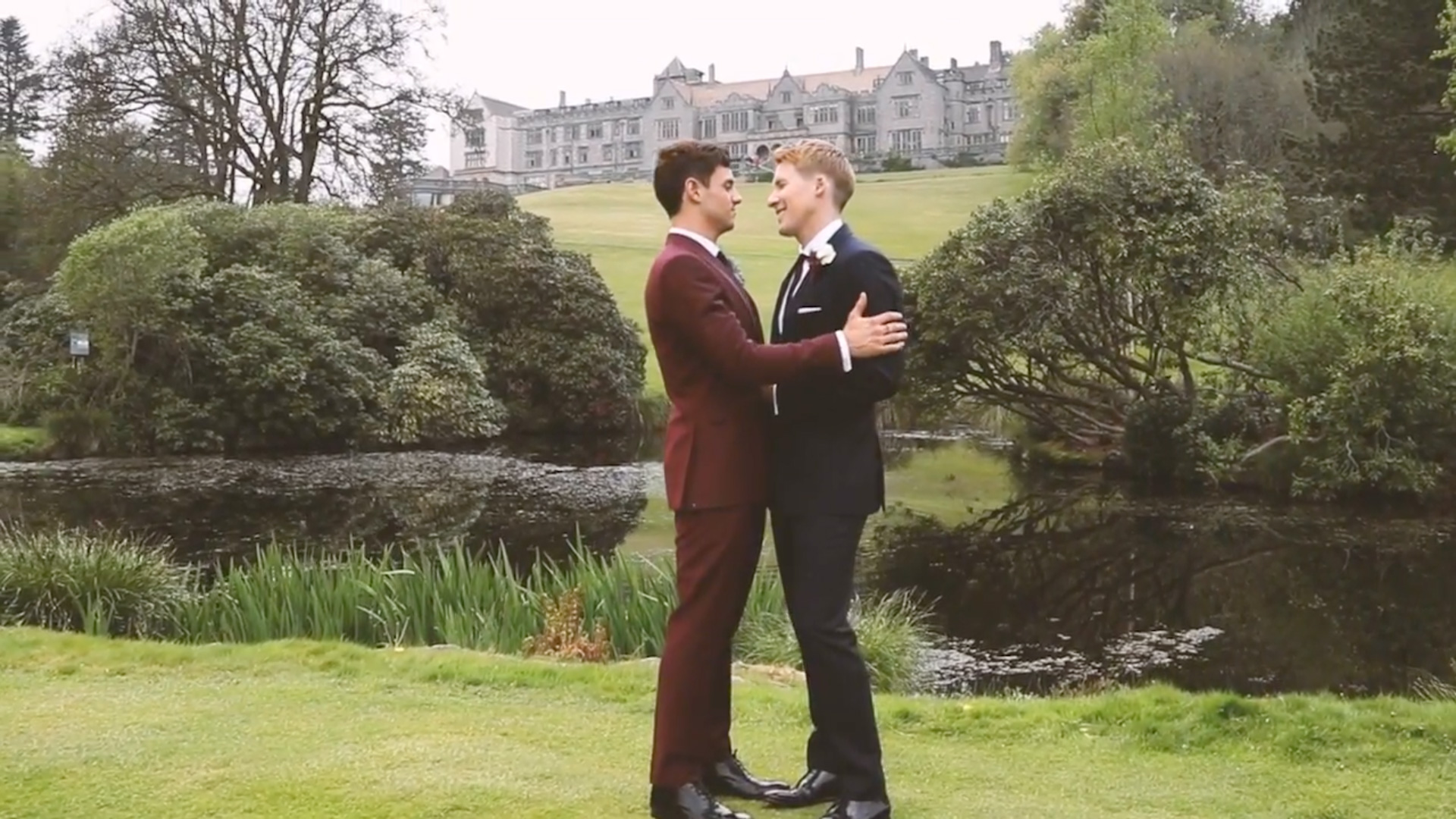 Tom Daley and Dustin Lance Black holding each other on their wedding day in suits infant of a castle and grass and a lake