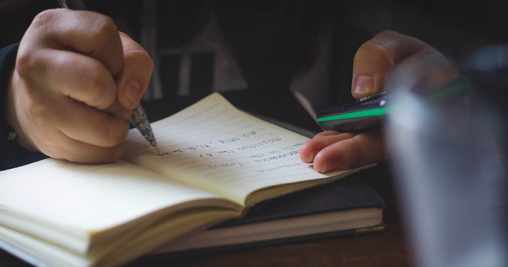 A person writing in a notebook and holding a phone to symbolise that the majority of lgbt+ youth in Northern Ireland feel unwelcome in school