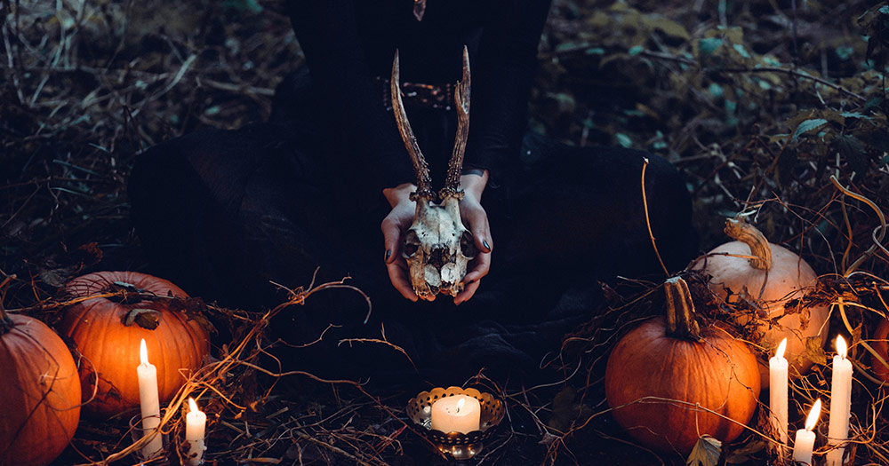 somebody hold a skull with horns in their hands while sitting in front of pumpkins and candles in the woods