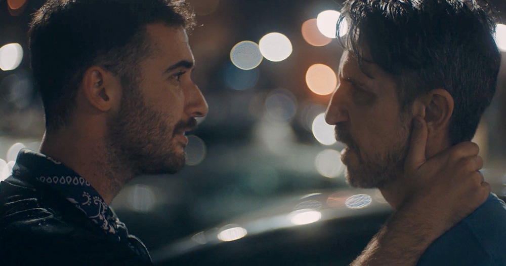 A still from the film call your father with two men looking at each other intensely at night with lights in the background