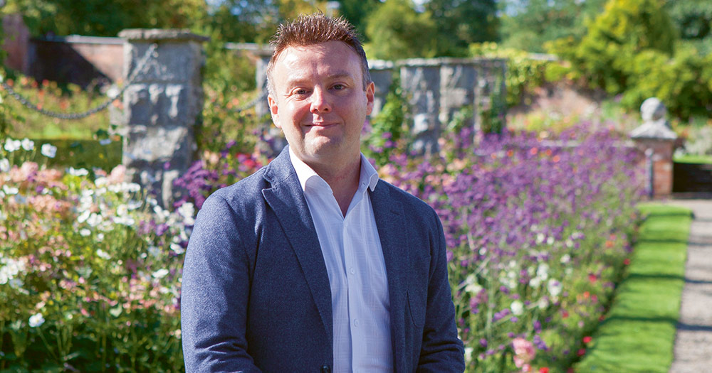 Daragh Doyle, the wedding planner from Rainbow Weddings, wearing a blue suit jacket in front of gorgeous purple flowers and a green lawn