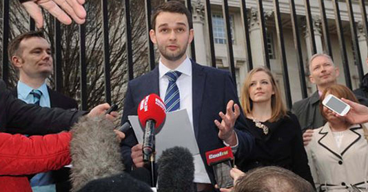 A man stands talking to a crowd of reporters, gesturing with his left hand.