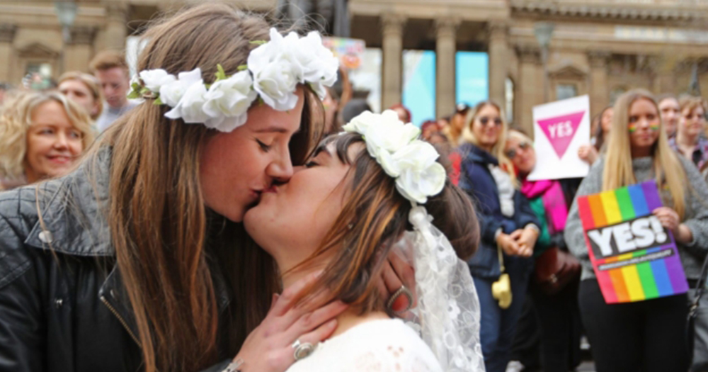 Two women in wedding dresses kiss with marriage equality posters in background