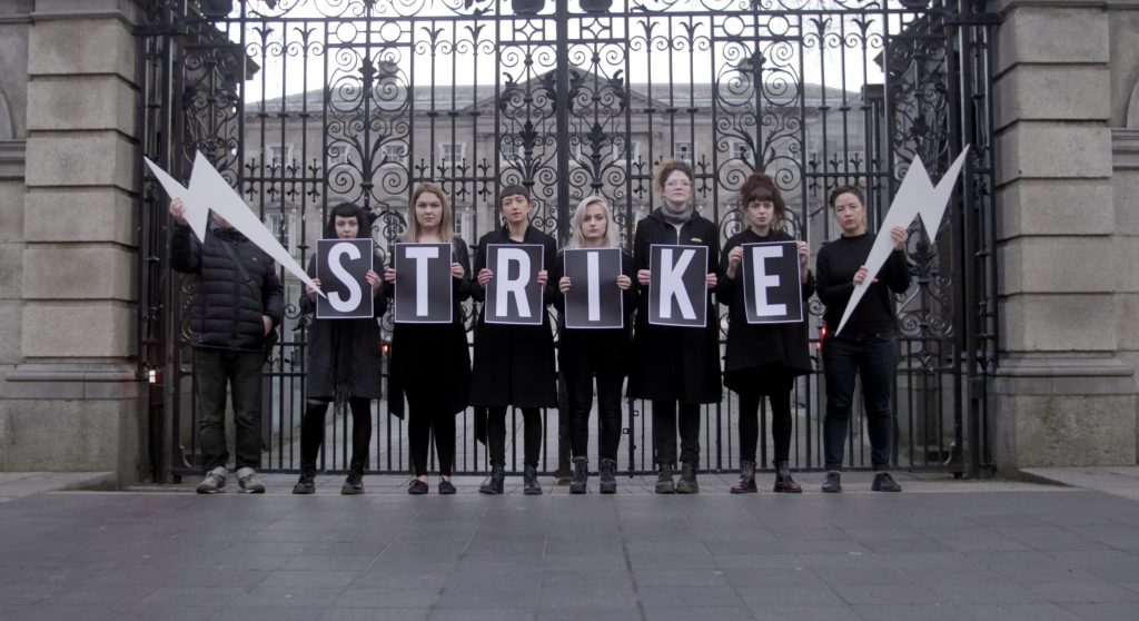 Image of woomen dressed in black standing outside leinster house with a strike banner