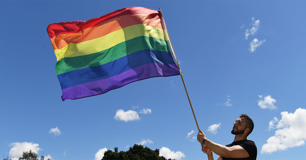 New Zealand man waves a pride flag against the sky as a backdrop