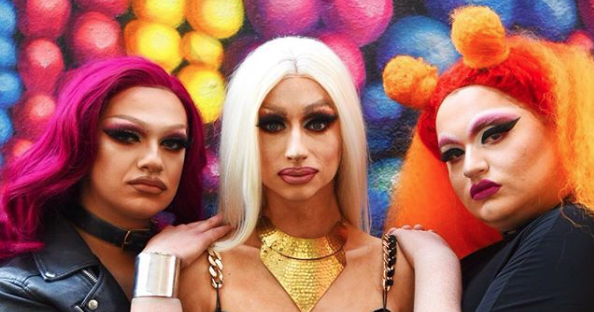 Three drag queens in full makeup posing against a multi coloured wall