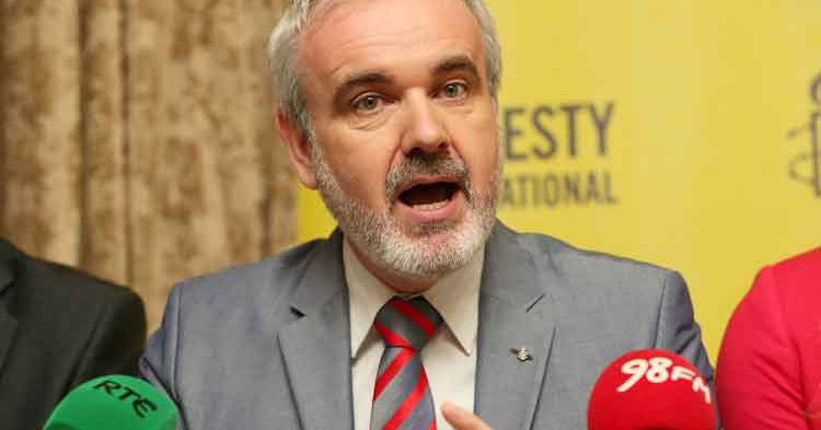 Colm O'Gorman at a press conference speaking into a microphone with the Amnesty International logo behind him