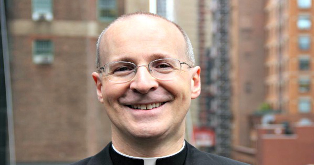 Father James Martin, the subject of a petition to block him from the Papal visit event, stands smiling with a city scape in the background