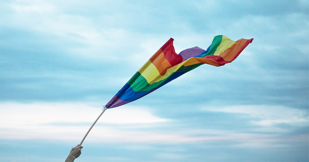 A hand holds the Pride flag blowing the wind against the sky at Belfast Pride
