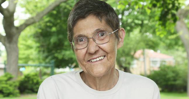 Elderly lesbian woman Marsha Wetzel sits on a bench in front of trees dressed in a light jumper and glasses smiling at the camera