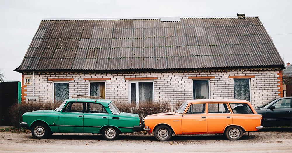 Two ancient looking cars parked bumper to bumper outside a small house in a Russian village