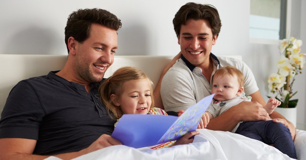 Male gay parents sit in bed with their two small children from adoption reading a book