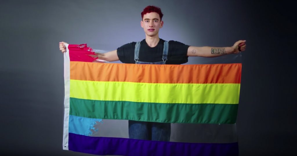 Image of Olly Alexander holding Pride Flag with the red and blue stripes ripped out.