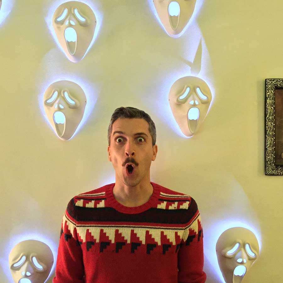 A man at a Halloween party, behind him lit up scream masks on a wall