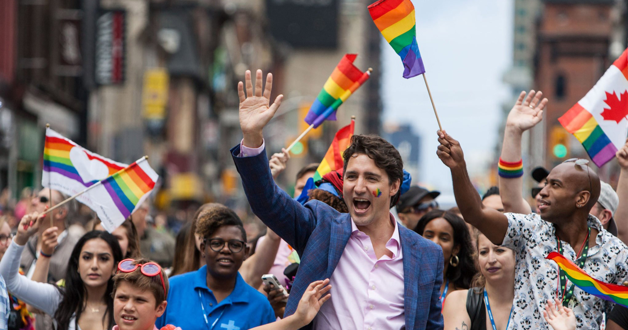 canadian Prime Minister Justin Trudeau at Montreal Pride with a rainbow painted on his cheek, waving at the crowds as he marches.