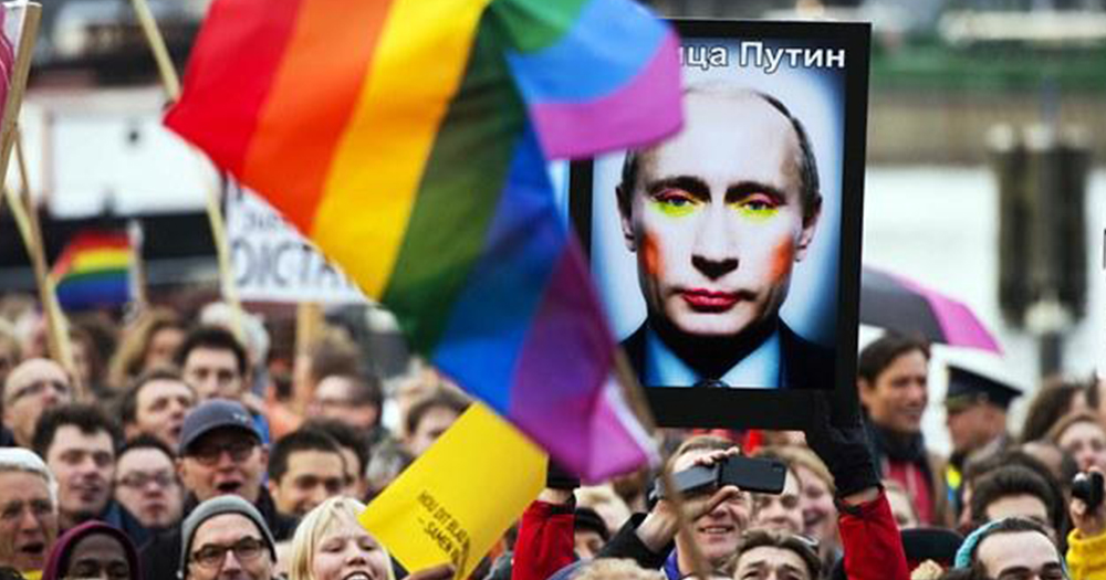 A crowd of LGBT+ supporters are gathered, a rainbow flag is in the foreground and a poster of Russian leader Putin wearing make-up is seen in the background