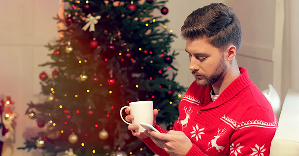 Tips to look after your mental health this Christmas - a young man drinking from a cup while checking his phone. A Christmas tree is in the background