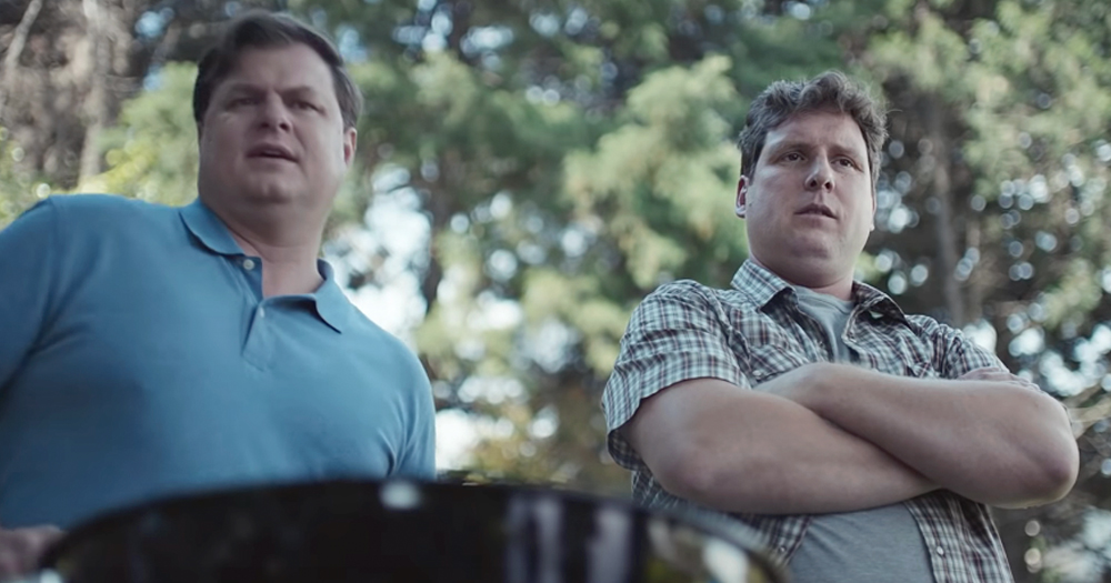 Still from new Gillette ad showing two men at a barbecue, one in a blue shirt and one in a checkered shirt with his arms crossed