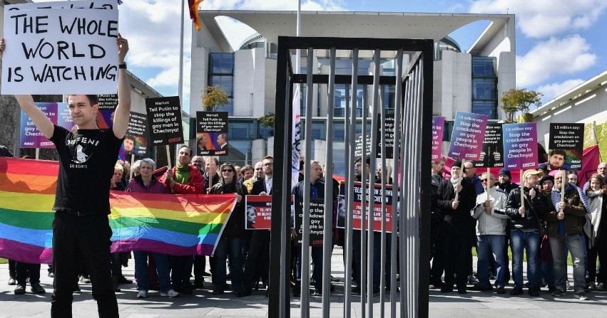 A group of protestors hold signs, rainbow flags in front of a fake prison cell, protesting the purge of LGBT+ people in Chechnya.