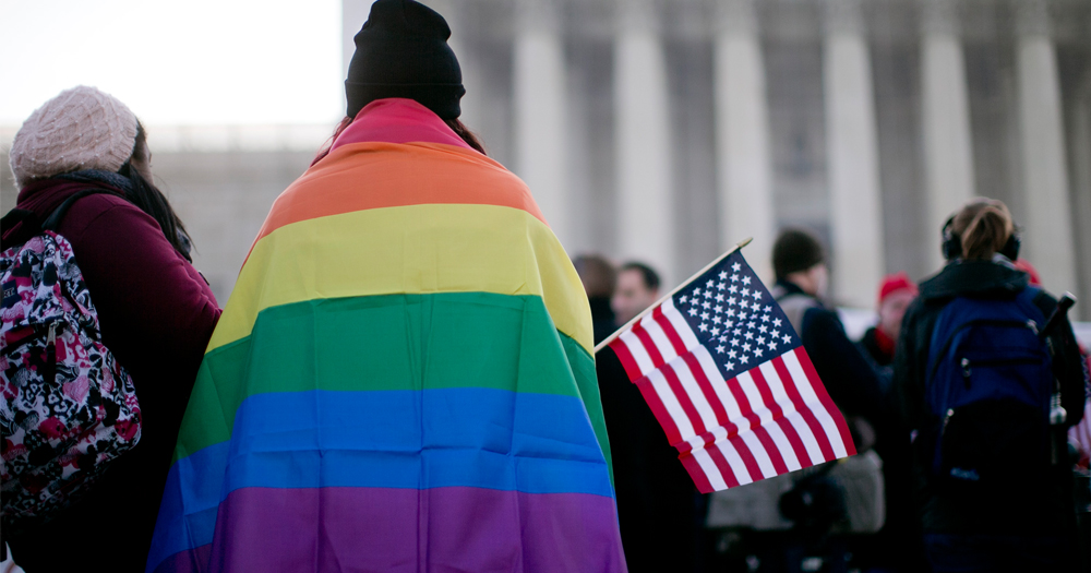 Person in Washington DC with a rainbow flag over their back, holding an American flag