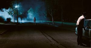 A man locks a car door while in the background a naked woman watches in a still from the Kate Dolan film