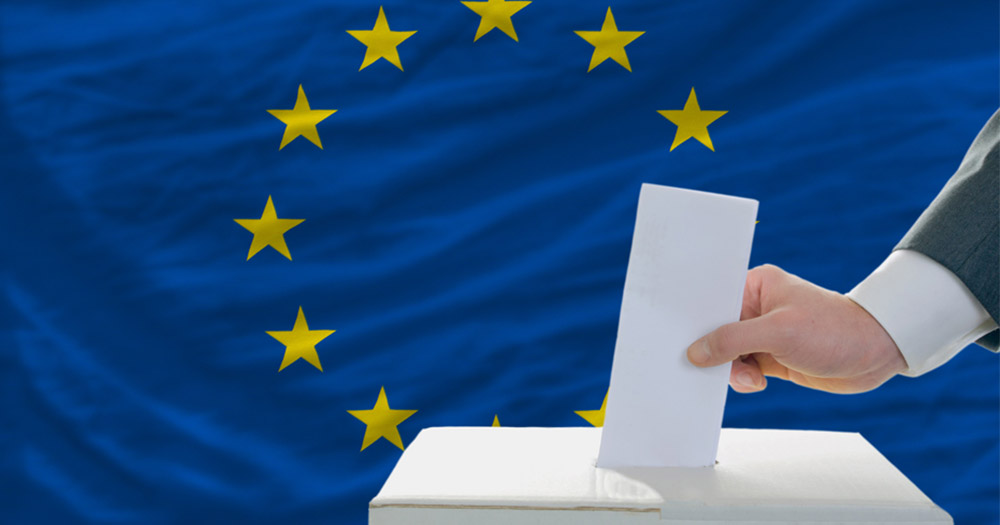 A hand places a paper vote into a ballot box backed by the European flag