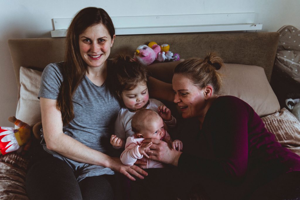 Female same sex parents on the couch at home, their toddler daughter in the middle holding her newborn sister