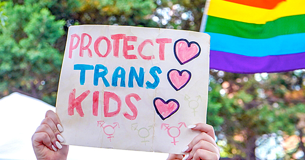 A banner that reads 'Protect Trans Kids' and a rainbow flag in the background.