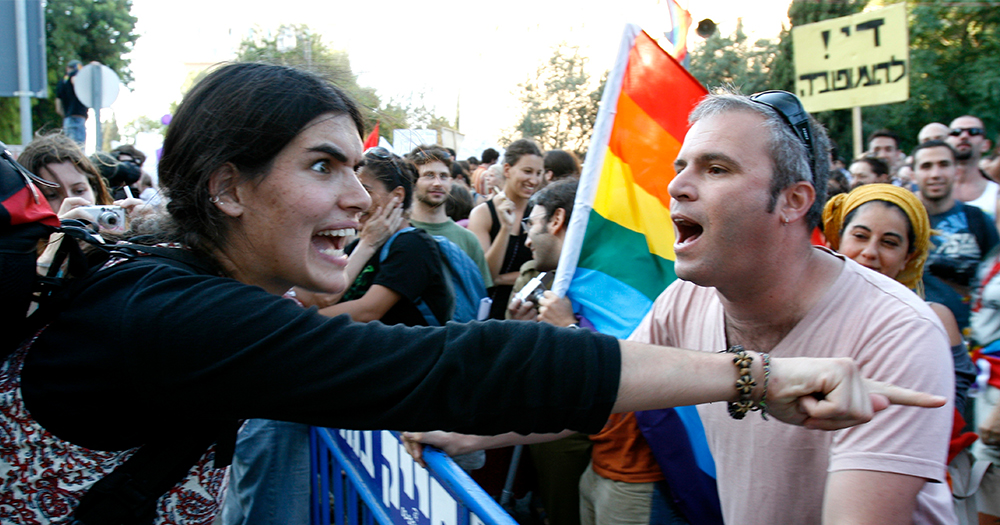 An irate woman screams with pointed finger at LGBT+ people marching in a gay pride parade holding rainbow flags protesting homophobic attacks