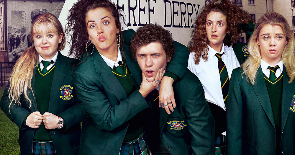 The cast of Derry Girls, four young women and one young man pose comically in front of a Free Derry sign