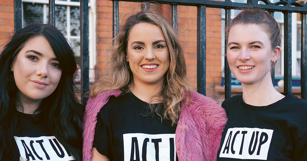 Three women wearing ACT UP t-shirts standing beside a fence