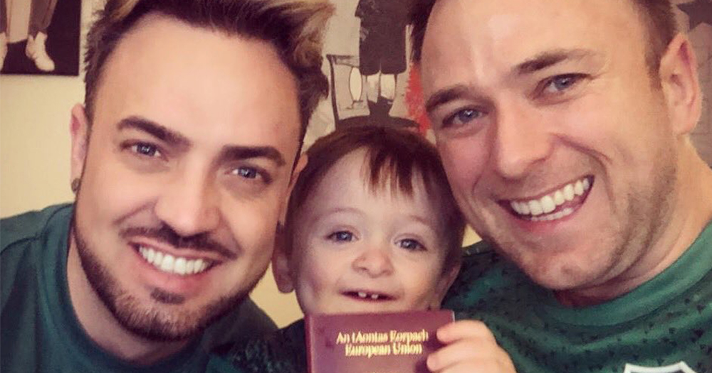 Irish gay dads pose with their toddler son in the middle holding a passport