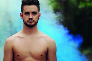 A topless bearded young man stands by a river