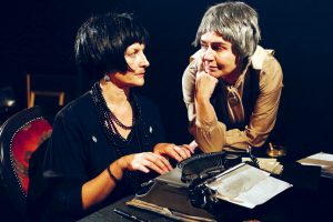 Two middle aged women wearing wigs lean over a typewriter