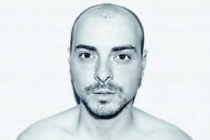 A headshot of a bearded shaven headed topless man