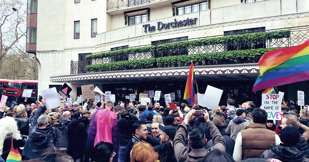 A group of protestors outside The Dorchester Hotel holding rainbow flags