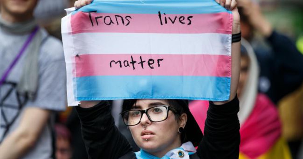 Person holding up a trans flag reading 'trans lives matter' at a protest against transphobic assault