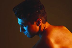 A topless young man lit by a phone screen