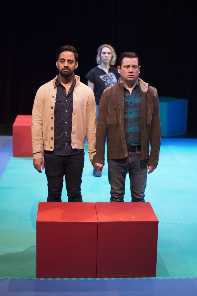 Two men standing beside each other holding hands. A woman behind them in the distance looking at them. This is a moment from the show The Baby Monitor