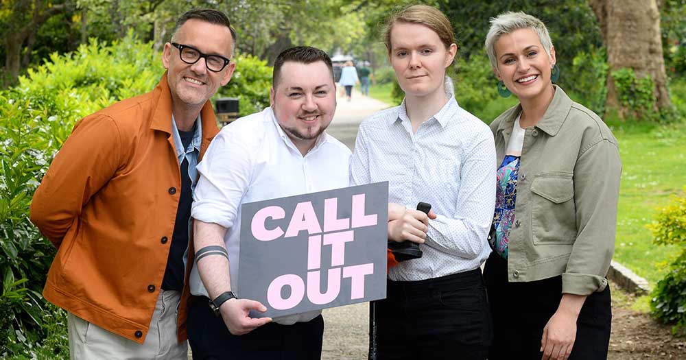 Four people hold a Call It Out' sign. They are a man, a woman, a trans man and a trans woman. they are standing in a park.